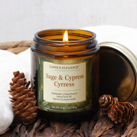 A lit amber glass candle jar labeled Candle Elegance Sage & Cypress Cypress, styled with pinecones and dried leaves on a rustic surface with a white cloth and metal lid beside it.