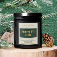 A black glass jar candle with a black lid and a cream label reading 'Candle Elegance' and 'Fraser Fir Noir,' styled on a wood slice with evergreen branches, burlap, and a pinecone against a green textured backdrop.