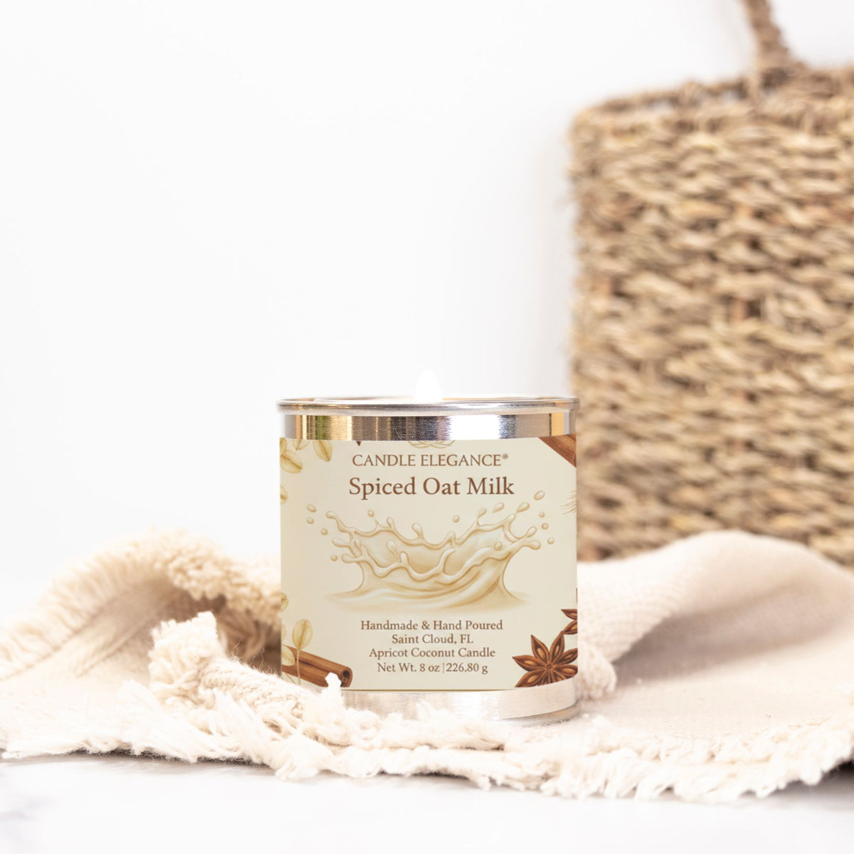 A Candle Elegance 'Spiced Oat Milk' handcrafted coconut candle in a silver tin, styled on a cream fringed cloth with a woven basket in the background.