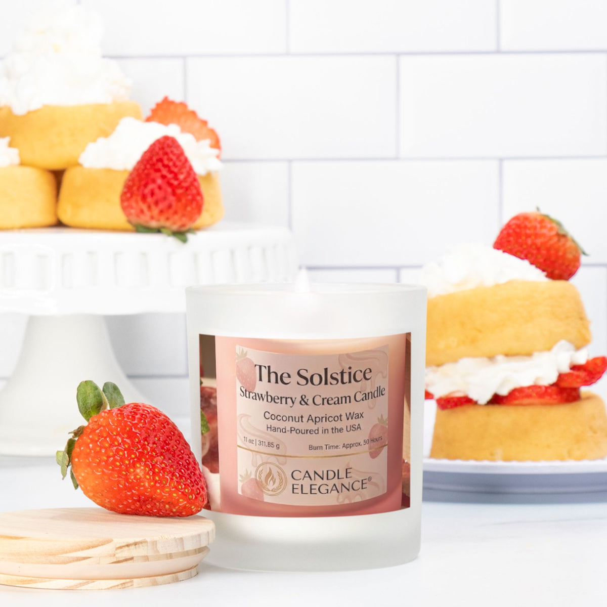A frosted glass candle jar labeled 'The Solstice Strawberry & Cream Candle' by Candle Elegance, styled on a white surface with a blurred kitchen scene of strawberries and cream cakes in the background.