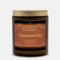A Candle Elegance Cinnamon Chai scented candle in an amber glass jar with a gold metal lid and a brown label with gold text, set against a light gray background.