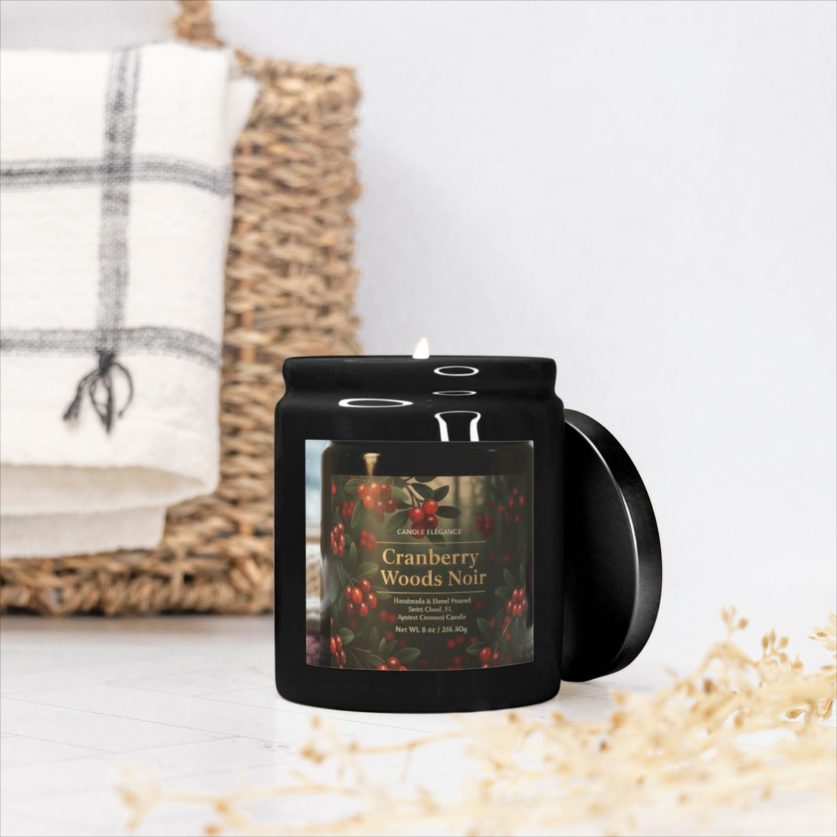 A black jar candle with a lit wick and a circular lid leaning against it, placed in front of a woven basket with a white plaid towel. The front label reads 'Cranberry Woods Noir' and shows cranberry illustrations.