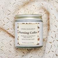 A glass jar candle labeled 'Candle Elegance Morning Coffee' sits on a cream-colored textured knit blanket. The candle has a white label with brown text and coffee bean illustrations, a silver metal lid, and a clear glass container showing the wax inside.
