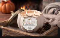 A hand-poured amber spice candle in a pumpkin-shaped jar with a gold lid resting beside it on a wooden tray. The candle is lit with two flames, decorated with a jute twine bow and a round paper label.