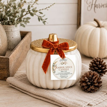 A white ribbed pumpkin-shaped candle jar with a gold lid and a rust-orange bow, set on a beige cloth with fall decor including pinecones, a small pumpkin, and a wooden tray in the background.