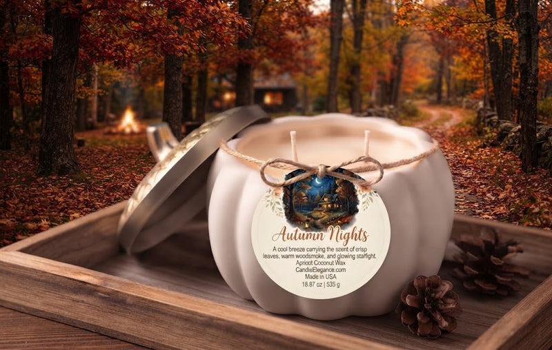 A white decorative pumpkin-shaped candle with its lid leaning against it, placed on a wooden tray in an autumn forest setting. The candle has a round paper label reading 'Autumn Nights' and is styled with pinecones and a small campfire scene in the background.