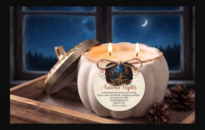 A white pumpkin-shaped candle with a gold-toned lid resting beside it on a wooden tray, lit with two flames. The candle has a round paper label reading 'Autumn Nights' and is styled with pinecones in front of a window showing a night sky and moon.