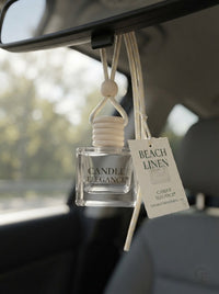 A clear glass car air freshener bottle labeled 'CANDLE ELEGANCE' hanging from a car rearview mirror, with a cream-colored reed diffuser top and a hanging tag that reads 'BEACH LINEN'.