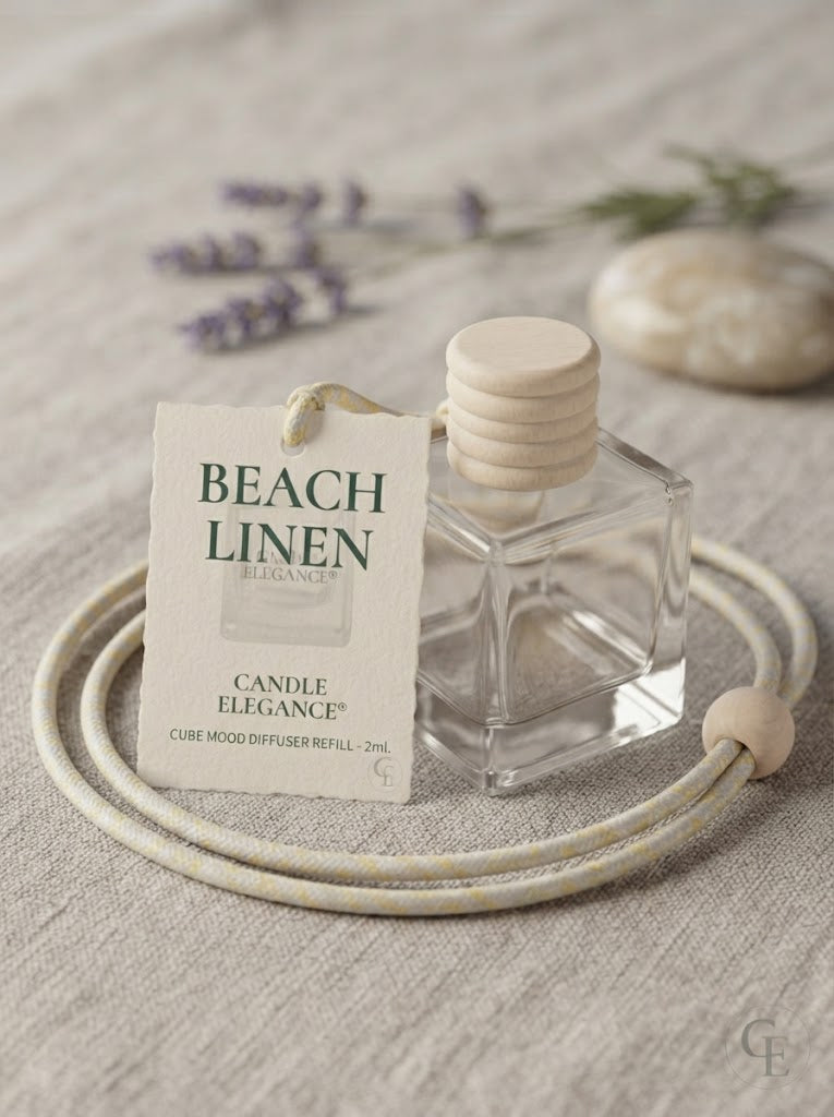 A clear glass cube-shaped candle diffuser bottle with a light wood cap sits beside a cream-colored coiled cord and a printed tag reading 'BEACH LINEN' and 'CANDLE ELEGANCE,' on a textured beige fabric surface with soft blurred lavender and stone props in the background.