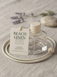 A clear glass cube-shaped candle diffuser bottle with a light wood cap sits beside a cream-colored coiled cord and a printed tag reading 'BEACH LINEN' and 'CANDLE ELEGANCE,' on a textured beige fabric surface with soft blurred lavender and stone props in the background.