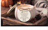 A pumpkin-shaped candle with its lid leaning beside it on a rustic wooden tray. The candle is wrapped with a twine bow and has a round paper label that reads 'CASHMERE VANILLA CANDLE.
