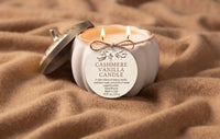 A lit double-wick candle in a white ceramic jar with a silver lid resting beside it on a soft brown fabric background. The front label reads 'CASHMERE VANILLA CANDLE' with a small decorative illustration above the text.