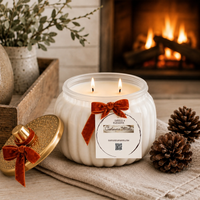 A Candle Elegance white ribbed glass jar candle with two lit wicks, tied with a rust-brown velvet bow and a white product label, placed on a beige cloth beside a gold lid. A cozy fireplace with logs burns in the background, with pinecones and a rustic tray nearby.
