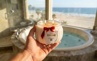 A hand holds a Candle Elegance double-wick candle in a clear glass jar with cream-colored wax and a red ribbon bow, set against a blurred background of a beach and a hot tub in a bathroom.
