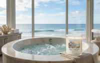 A Coastal Cottage Candle in a clear glass jar sits on a beige towel beside a marble hot tub with ocean water and a beach view through large floor-to-ceiling windows.