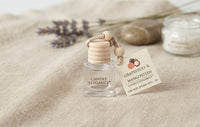 A small clear glass candle bottle labeled 'CANDLE ELEGANCE' with a light wood bead cap and a hanging tag reading 'GRAPEFRUIT & MANGOSTEEN'. The bottle is styled on a beige fabric surface with dried lavender, smooth stones, and a softly blurred jar in the background.