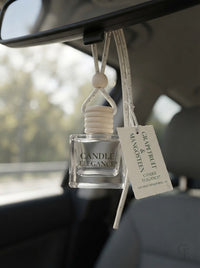 A clear glass car diffuser bottle labeled 'CANDLE ELEGANCE' hanging from a black car rearview mirror, with a white fabric reed diffuser top and a white hangtag attached. The diffuser is shown inside a car interior.