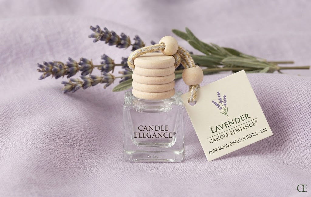 A lavender scented candle diffuser refill by Candle Elegance, featuring a clear glass bottle with a stacked light wood reed diffuser cap and a hanging paper tag, styled with dried lavender sprigs on a soft lavender fabric background.