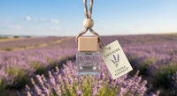 A small glass reed diffuser bottle with a wooden cap hangs from a braided cord with a wooden bead, set against a blurred lavender field. A paper tag reads 'LAVENDER' and 'CANDLE ELEGANCE'.
