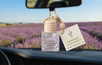 A lavender scented car diffuser bottle with a wooden bead top and a hanging tag, displayed on a car dashboard with a blurred lavender field in the background.