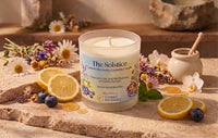 A scented candle in a clear glass jar labeled 'The Solstice' by Grommet Fragrance, styled on a stone surface with lemon slices, blueberries, dried flowers, and a small ceramic pot with a wooden spoon.