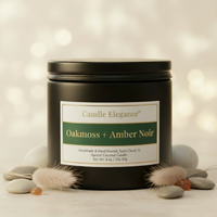 A matte black metal candle tin with a cream and green label reading 'Candle Elegance Oakmoss + Amber Noir,' styled with faux fur and smooth stones on a warm beige surface against a softly blurred bokeh background.