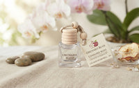 Orchard Bliss Canary Elegance diffuser refill bottle with a wooden bead cap, shown next to its product tag on a beige linen surface with spa stones, dried apple slices, and soft orchid flowers in the background.