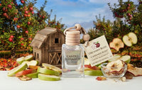 A clear glass perfume bottle labeled Candle Elegance with a wooden stopper and hanging tag, set on a table with sliced apples, dried apple pieces, and an orchid blossom illustration. A rustic barn and apple trees appear in the blurred background.