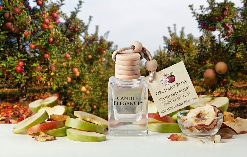 A clear glass perfume bottle labeled Candle Elegance stands on a white surface with apple slices, dried apple pieces, and a hanging tag reading Orchard Bliss by Candle Bliss. The background shows an apple orchard with red and green apples among trees.