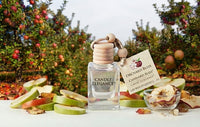 A clear glass perfume bottle labeled Candle Elegance stands on a white surface with apple slices, dried apple pieces, and a hanging tag reading Orchard Bliss by Candle Bliss. The background shows an apple orchard with red and green apples among trees.
