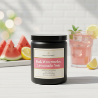 A black candle jar labeled 'Pink Watermelon Lemonade Noir' by Candle Elegance, set on a white surface with watermelon slices, a pink drink, and lemon slices in a bright, softly blurred background.