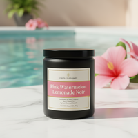 A black jar candle labeled 'Candle Elegance' in 'Pink Watermelon Lemonade Noir' scent, placed on a white marble surface with a blurred swimming pool and pink flowers in the background.