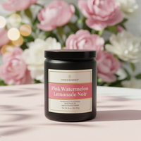 A black candle jar labeled 'Candle Elegance' and 'Pink Watermelon Lemonade Noir' sits on a light pink surface with a blurred background of pink and white flowers.