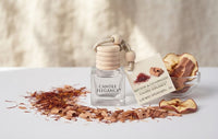 A clear glass diffuser bottle labeled CANDLE ELEGANCE stands on a white surface with dried saffron threads scattered around it. A small card attached to the bottle reads Saffron & Cedarwood, and a glass bowl containing dried apple slices sits to the right against a softly blurred neutral background.