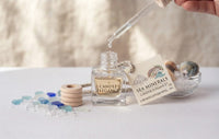 A clear glass bottle labeled 'Candle Elegance' with a glass dropper above it, alongside a card reading 'Sea Minerals' and decorative stones and a small bowl on a white surface.