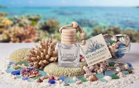 Sea Minerals Cube Mood Diffuser by Candle Elegance featuring a clear glass bottle with a wooden lid, surrounded by coral, seashells, and sea glass on a sandy background.