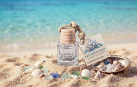 Sea Minerals Cube Mood Diffuser by Candle Elegance featuring a clear glass bottle with a wooden lid, wooden cord, and a labeled retail card, displayed on a sandy beach setting with seashells and sea glass.