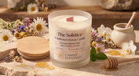 A frosted glass candle jar labeled 'The Solstice Wildflower Honey Candle' by Candle Elegance, shown with a pink wooden wick and a wooden lid beside it on a rustic stone surface. The scene includes dried flowers, mint leaves, honey, and a small ceramic jar in a warm, styled setting.