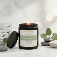 A lit black glass jar candle labeled 'Spa Day Noir' by Candle Elegance, styled on a marble surface with rolled towels, eucalyptus leaves, and smooth stones in a spa-inspired setting.