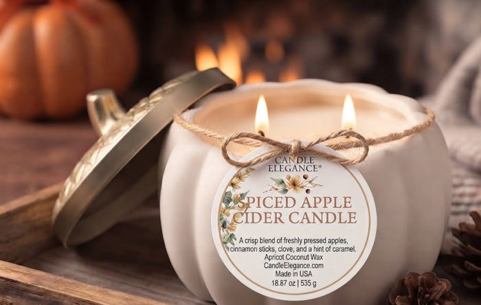 A lit spiced apple cider candle in a white pumpkin-shaped jar with a gold lid leaning beside it. The front label reads 'Candle Elegance' and 'Spiced Apple Cider Candle,' with twine tied around the jar.