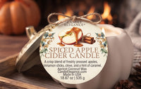A spiced apple cider candle by Candle Elegance, shown with its lid off and a round product tag tied with twine. The candle is in a white vessel on a rustic wooden surface with a blurred pumpkin and warm fire in the background, indicating a fall/spiced cider scent.