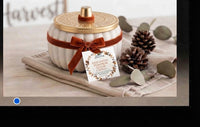 A white ribbed ceramic jar candle with a gold lid and a brown ribbon bow, displayed on a beige cloth with pinecones and eucalyptus leaves. A product tag is attached to the front.