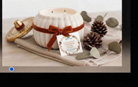 A white ribbed ceramic candle jar with a brown ribbon and bow, shown with its lid off and a product tag hanging from the front. The candle is styled on a beige cloth with pinecones, eucalyptus leaves, and a gold-toned decorative object on a light wood surface.