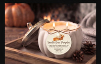 A white pumpkin-shaped candle with a metal lid leaning beside it, lit with two flames. The front label reads 'Vanilla Bean Pumpkin' with a pumpkin pie illustration and scent description.