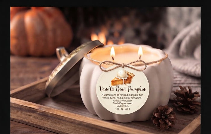 A white pumpkin-shaped candle with a metal lid leaning beside it, lit with two flames. The front label reads 'Vanilla Bean Pumpkin' with a pumpkin pie illustration and scent description.