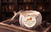 A pumpkin-shaped candle with its lid off, placed on a wooden tray in a cozy library setting with bookshelves and candles in the background. The candle has a round paper label that reads 'Vanilla Bean Pumpkin' and features a small illustration of a pumpkin and a slice of pie.