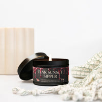 A candle in a black tin with the lid off, showing a lit wick. The label reads 'PINK SUNSET SIPPER' and features a dark design with red dripping graphics and small watermelon and cocktail illustrations.