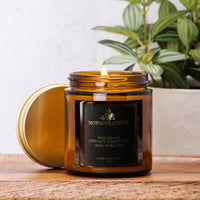 An amber glass jar candle with its lid leaning beside it on a wooden surface. The candle label reads 'MORNING COFFEE' and shows coconut apricot wax.