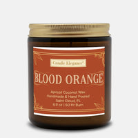 An amber glass jar candle with a gold screw-top lid and a terracotta-colored label reading 'Candle Elegance BLOOD ORANGE' on a light gray background.