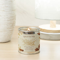 A lit Candle Elegance Spiced Oat Milk candle in a silver tin with a cream label, styled on a light wood surface next to a textured white vase and a soft-focus lamp.